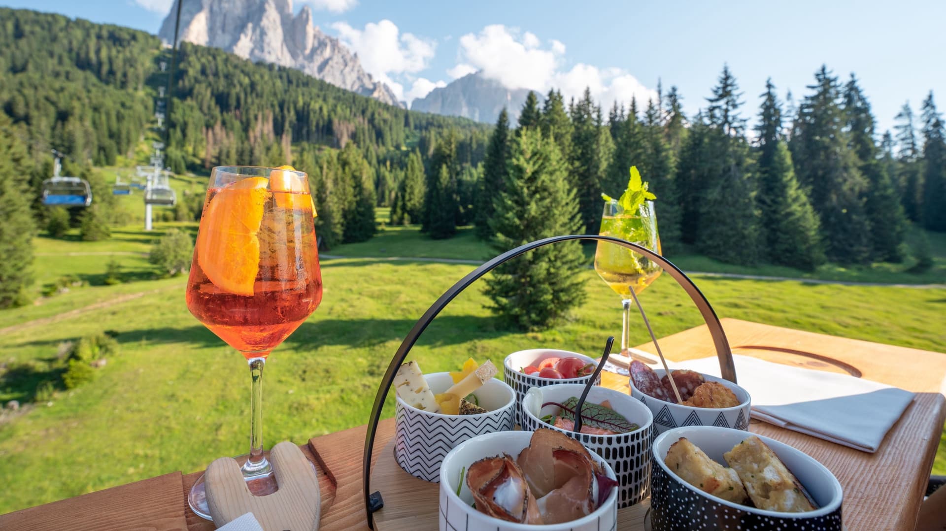 A tiered stand of antipasti and two glasses on the table, the chairlift rising between the firs toward the Sassolungo behind.