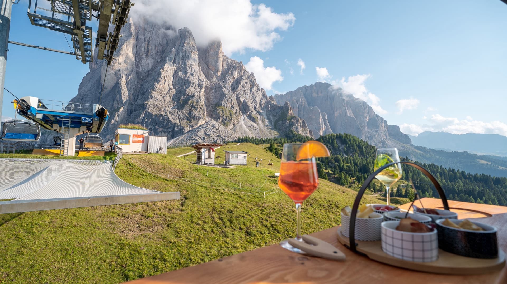 The Mont de Sëura chairlift station at the foot of the Sassolungo, two glasses and the antipasti baskets on the wooden table in the foreground.