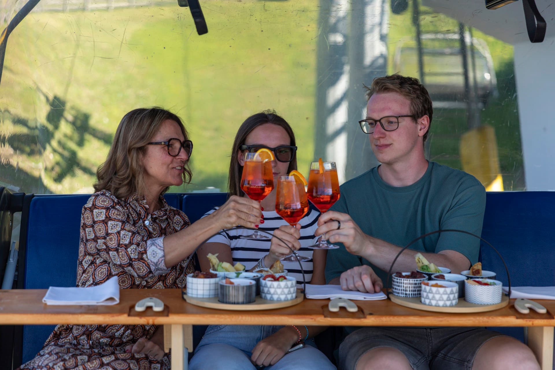 Three guests toasting with spritz on the chairlift, antipasti trays in front of them.